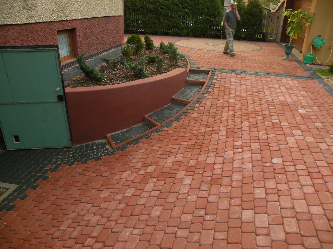 Czerwono-czarna kostka brukowa ułożona na podjeździe przed domem, z widocznym klombem obsadzonym iglakami i spacerującym mężczyzną w tle.