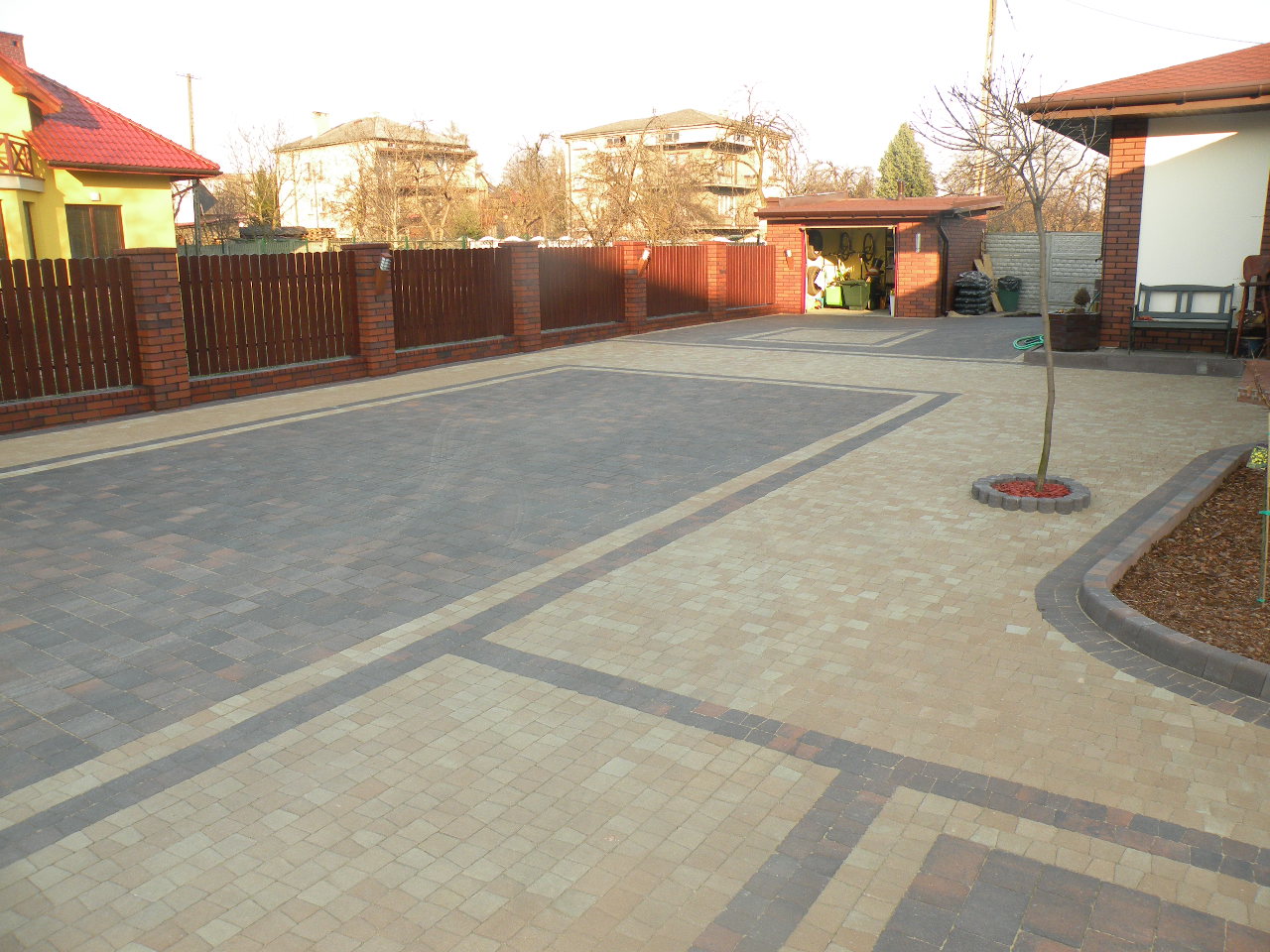 Ułożona kostka brukowa w dwóch odcieniach szarości i beżu tworząca wzór na podjeździe, z widocznym ogrodzeniem z cegły i drewna oraz garażem w tle.