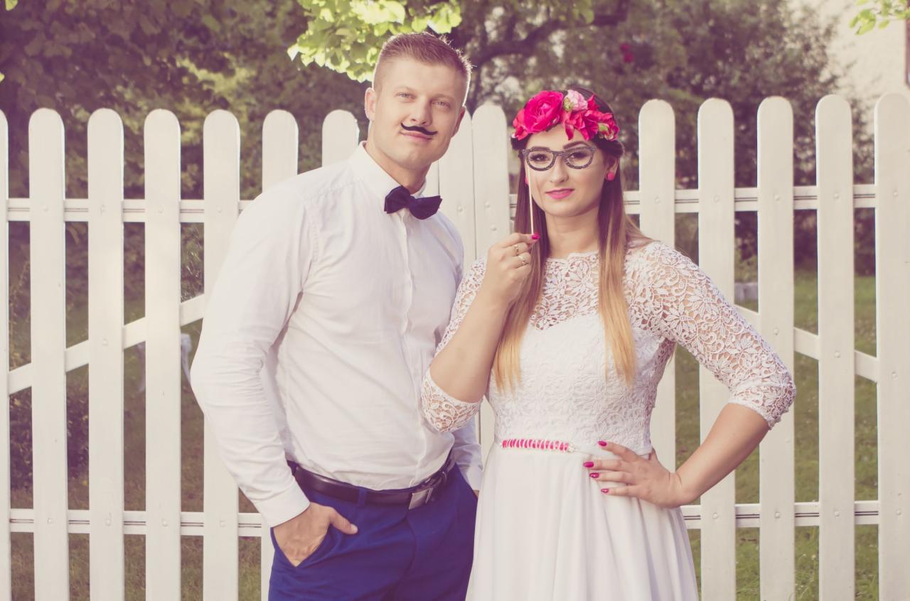 Para młoda w stylizacji retro z rekwizytami: wąsy i okulary na patyku, pozująca na tle białego płotu.