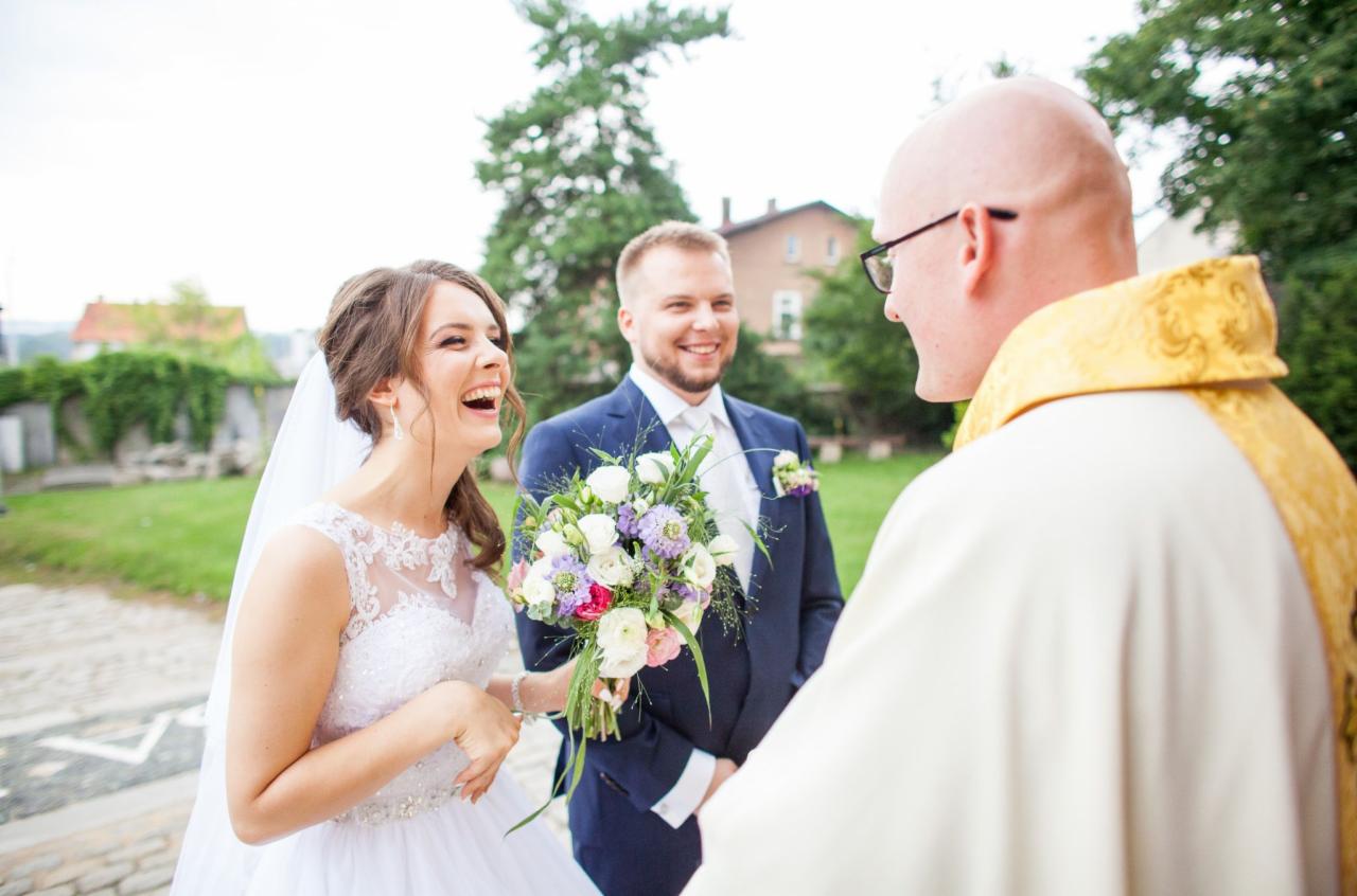 Uśmiechnięta panna młoda w białej sukni z koronkowym dekoltem i welonem, trzymająca bukiet kwiatów, rozmawia z księdzem podczas ceremonii ślubnej, w tle uśmiechnięty pan młody w granatowym garniturze.
