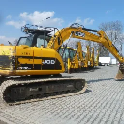 Żółta koparka gąsienicowa JCB JS130LC z widocznymi śladami użytkowania, zaparkowana na placu z kostki brukowej. W tle kilka innych maszyn budowlanych JCB oraz biały samochód dostawczy.