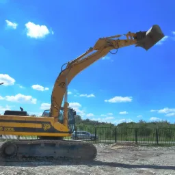 Żółta koparka JCB JS 330L z podniesionym ramieniem i łyżką na tle błękitnego nieba z obłokami, widoczna na placu budowy z ogrodzeniem i zielenią w tle.