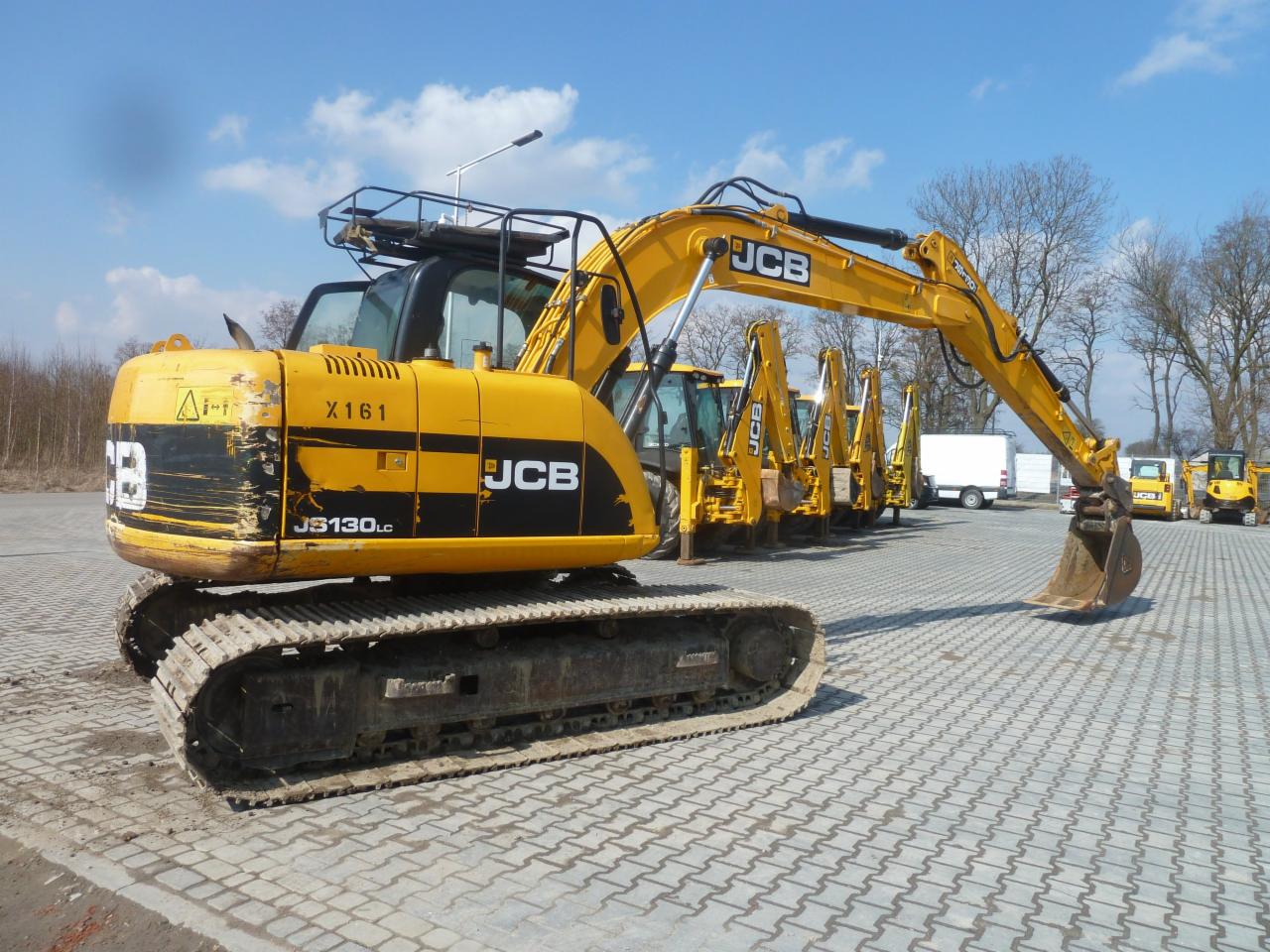 Żółta koparka gąsienicowa JCB JS130LC z widocznymi śladami użytkowania, zaparkowana na placu z kostki brukowej. W tle kilka innych maszyn budowlanych JCB oraz biały samochód dostawczy.