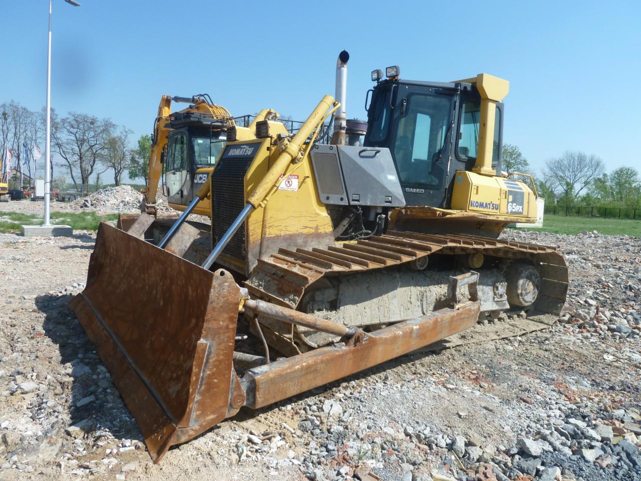 Żółty spychacz Komatsu 65PX z widocznymi śladami użytkowania na gąsienicach i lemieszu, zaparkowany na placu budowy z gruzem, w tle koparka JCB i drzewa pod błękitnym niebem.