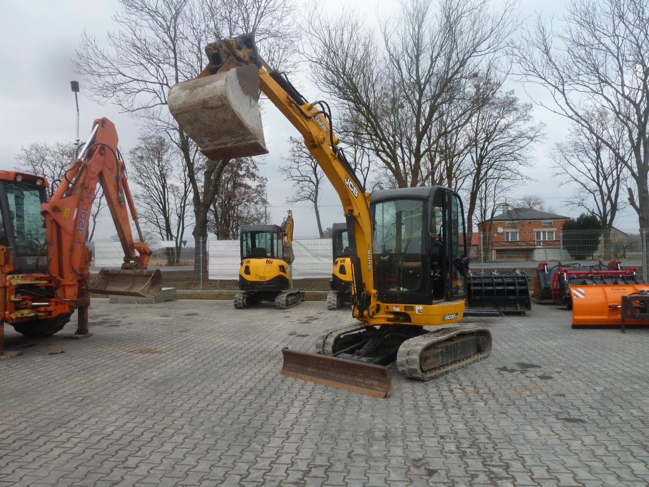 Żółto-czarna minikoparka JCB na placu z kostki brukowej, z podniesioną łyżką, w tle inne maszyny budowlane i budynek mieszkalny.
