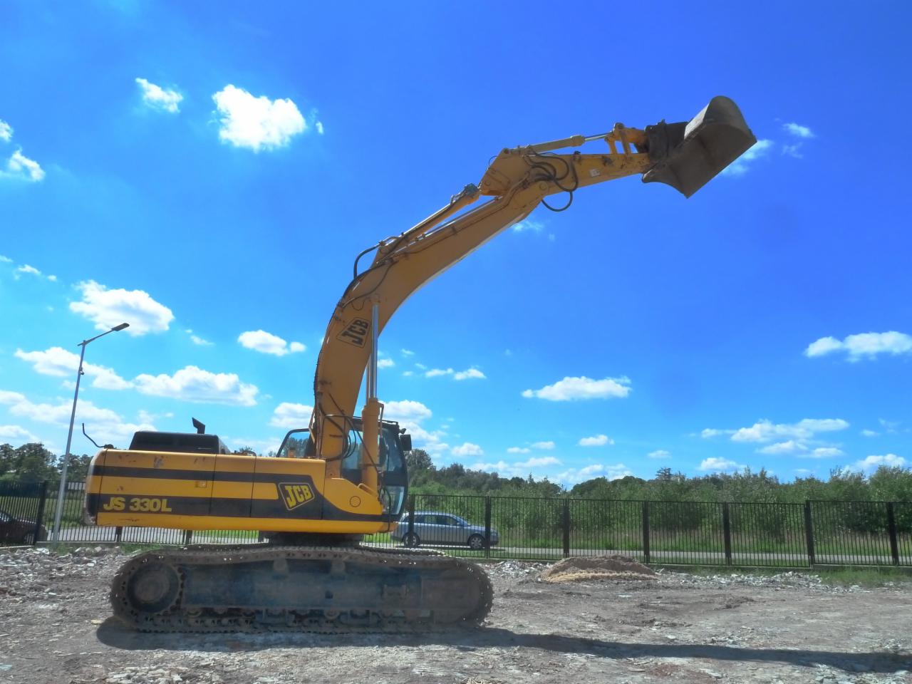 Żółta koparka JCB JS 330L z podniesionym ramieniem i łyżką na tle błękitnego nieba z obłokami, widoczna na placu budowy z ogrodzeniem i zielenią w tle.