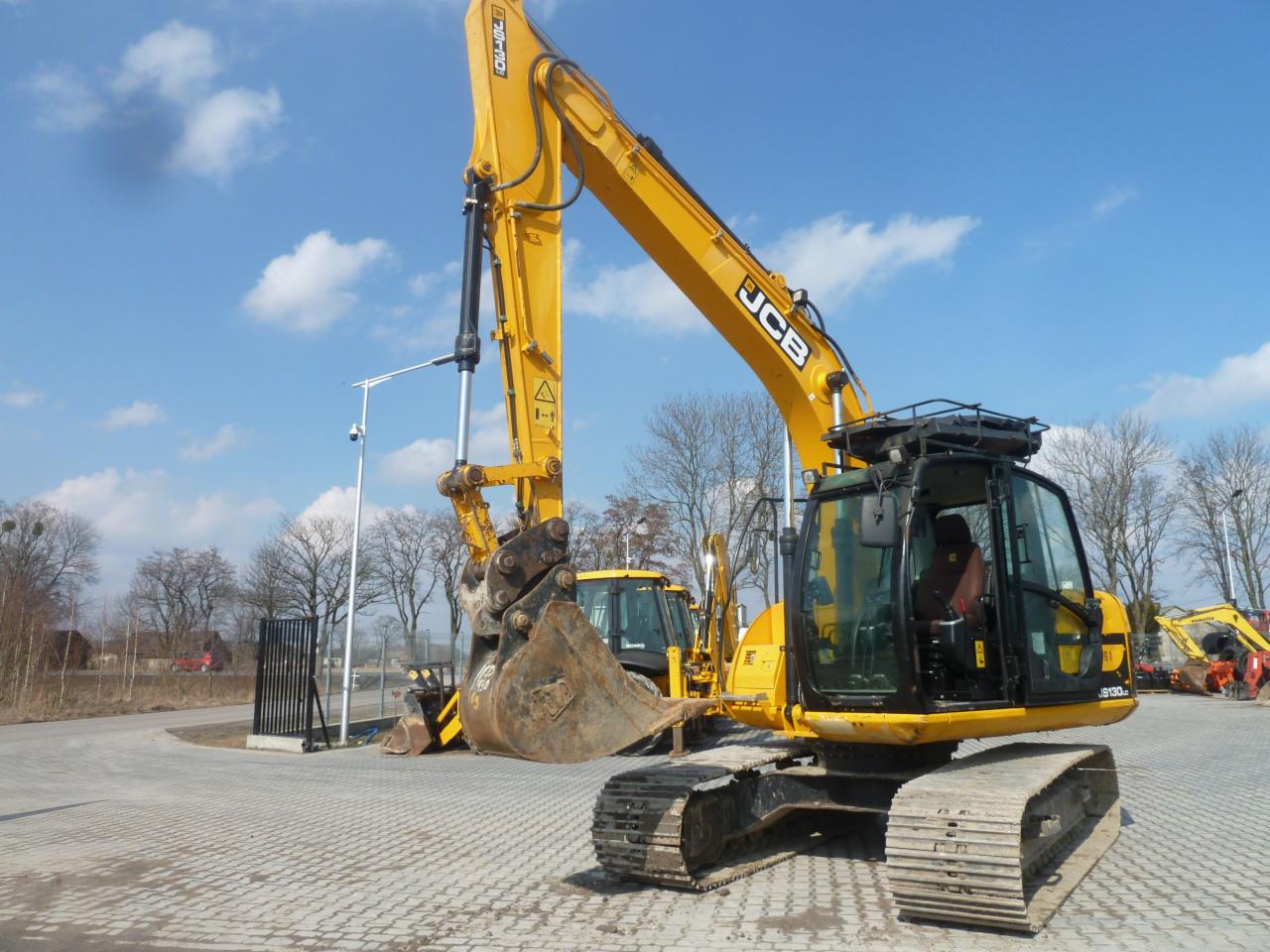 Żółta koparka gąsienicowa JCB 130 z łyżką, zaparkowana na placu wyłożonym kostką, z widocznymi innymi maszynami budowlanymi w tle i błękitnym niebem z chmurami.