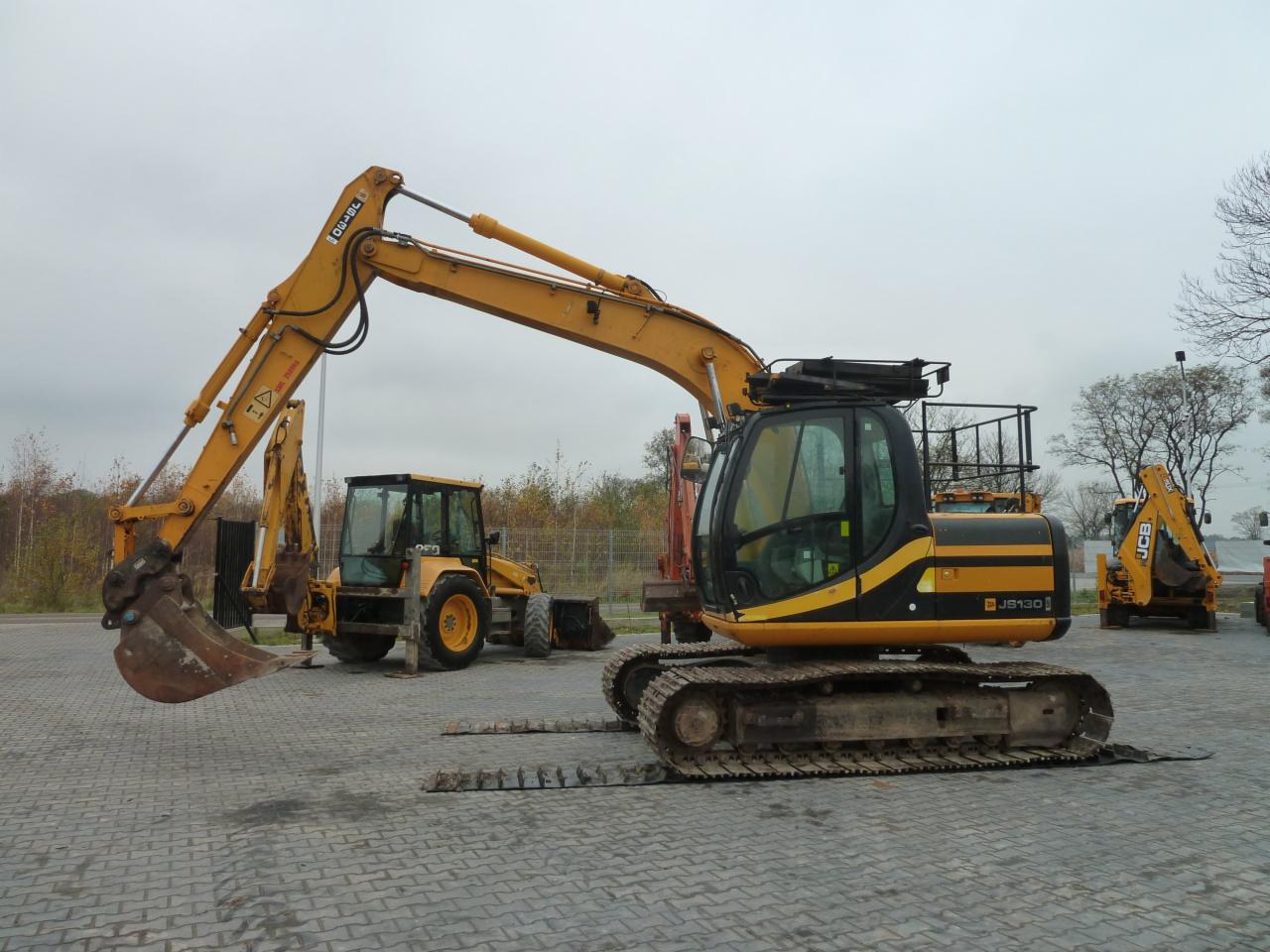 Żółto-czarny koparka gąsienicowa JCB JS130 na placu z kostki brukowej, w tle inne maszyny budowlane i drzewa bez liści pod pochmurnym niebem.