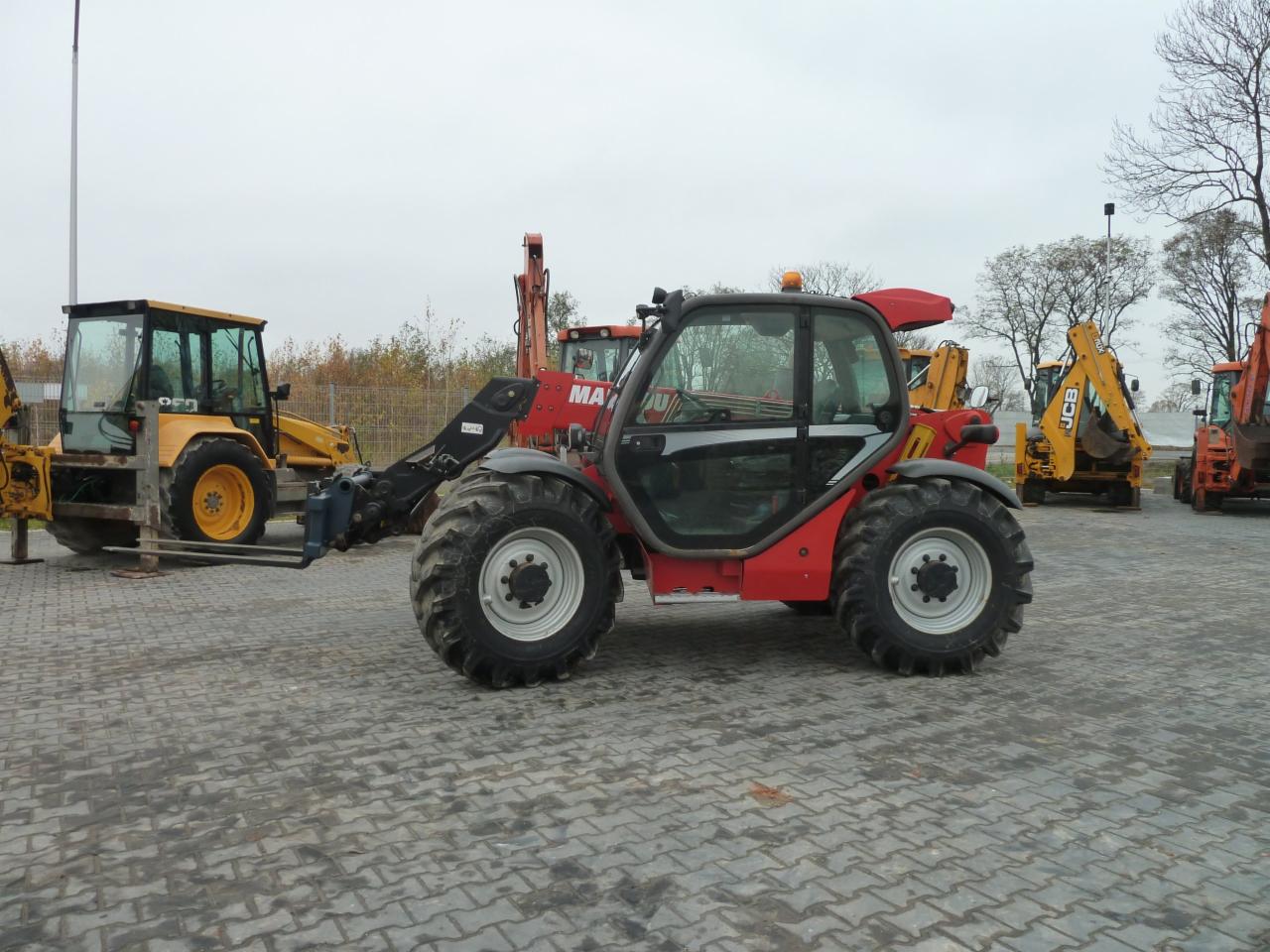 Czerwony teleskopowy wózek widłowy Manitou na placu z kostki brukowej, w tle żółte koparko-ładowarki JCB i inne maszyny budowlane.