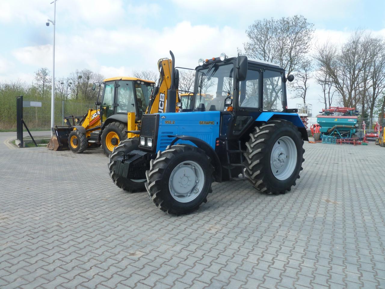Niebieski traktor marki Belarus 952.2 stoi na placu wyłożonym kostką, obok widoczny żółty traktor JCB z łyżką i wysięgnikiem, w tle drzewa i inne maszyny rolnicze.