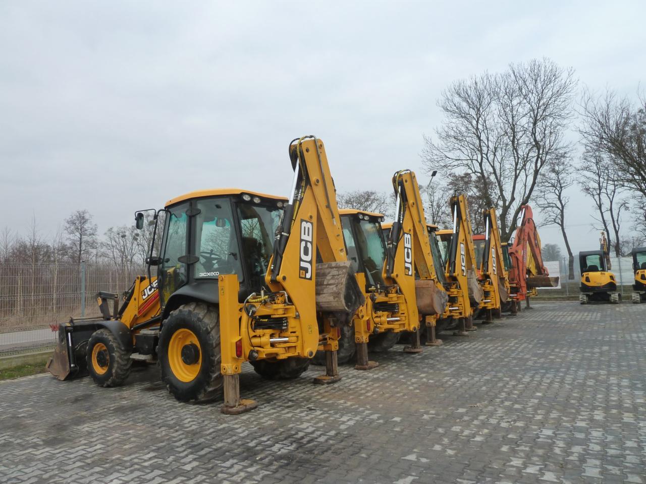 Rząd żółtych koparko-ładowarek JCB 3CX ECO zaparkowanych na brukowanym placu w pochmurny dzień. Widoczne podpory wysunięte, niektóre maszyny z łyżkami, inne z osprzętem kopiącym. W tle drzewa bez...