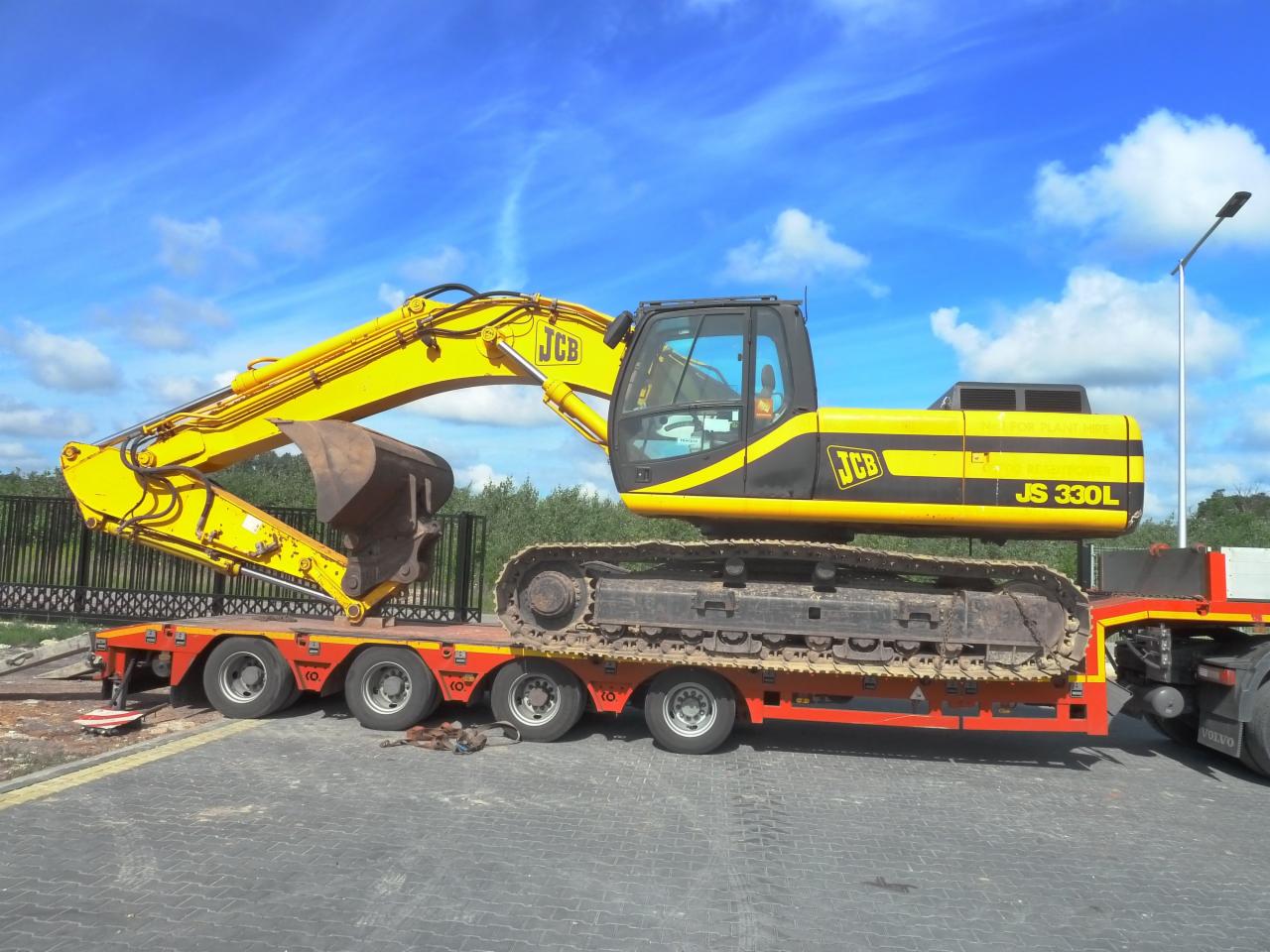 Żółta koparka gąsienicowa JCB JS 330L transportowana na niskopodwoziowej przyczepie z czerwonymi elementami, na tle błękitnego nieba z chmurami.