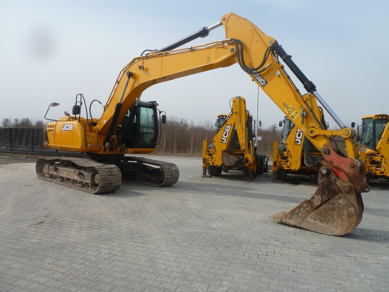 Żółta koparka gąsienicowa JCB na placu z kostki brukowej, z widocznymi innymi maszynami budowlanymi w tle, prezentowana z bliska z elementami roboczymi.