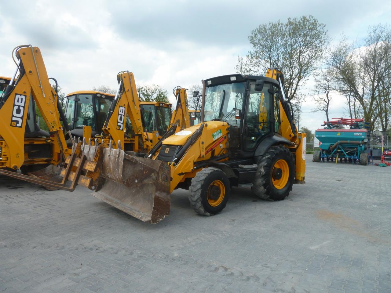 Żółte koparko-ładowarki JCB na placu, widoczna również maszyna rolnicza w tle.