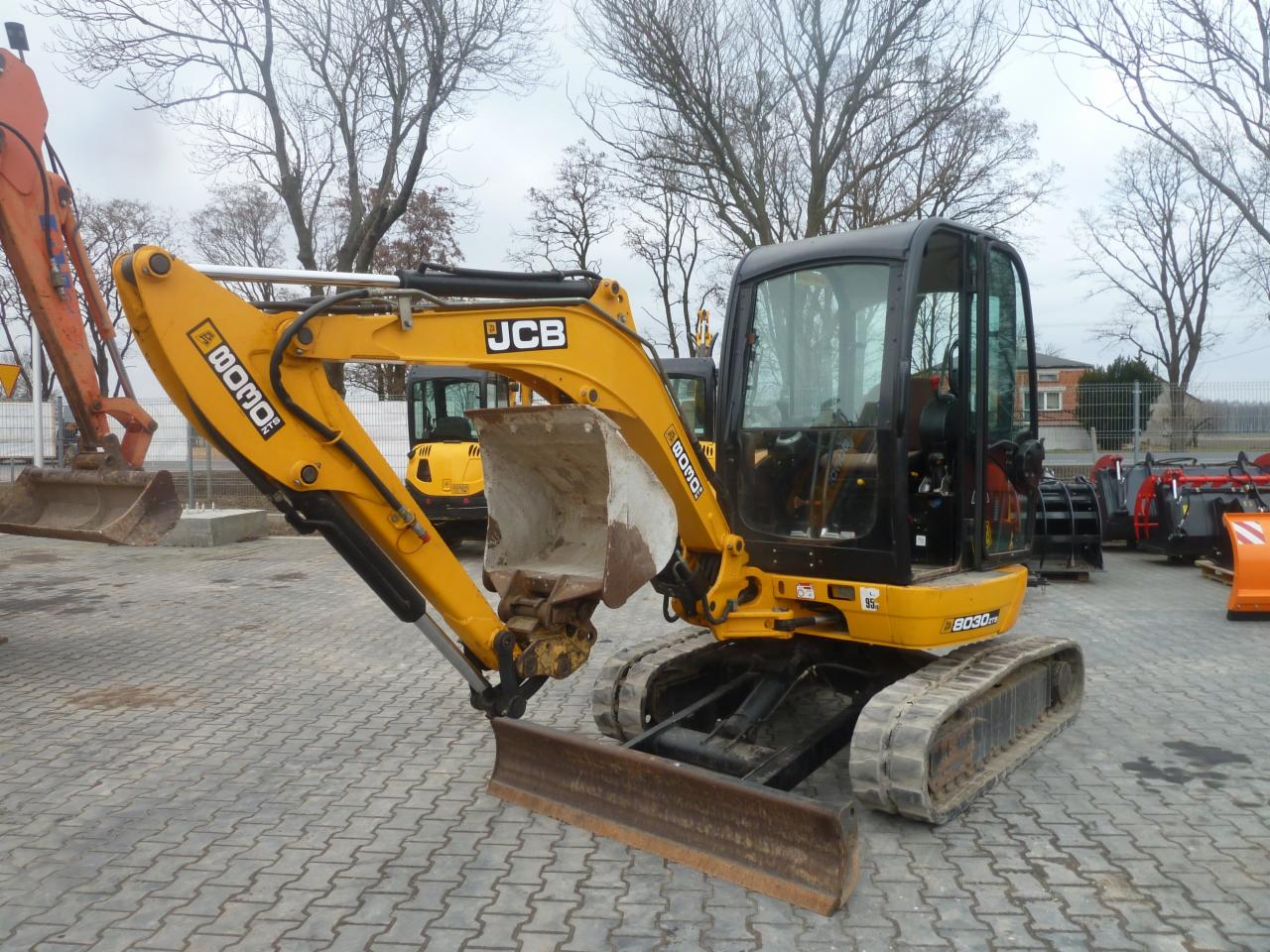 Żółta mini koparka JCB 8030 z łyżką i lemieszem stoi na brukowanym placu, widoczna z boku, z innymi maszynami w tle i nagimi drzewami na horyzoncie.