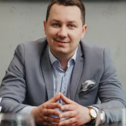 Portret biznesowy mężczyzny w szarej marynarce, siedzącego za stołem z rękami splecionymi na blacie, widoczny zegarek na ręce i poszetka w kieszonce marynarki.