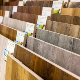 Ekspozycja próbek paneli podłogowych o różnych odcieniach drewna i fakturach, z etykietami producenta, ustawionych pod kątem na sklepowym stojaku.