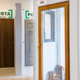Ekspozycja okna z drewnianą ramą w salonie sprzedaży, widoczne profile okienne wewnątrz, na drugim planie drzwi z logo producenta.