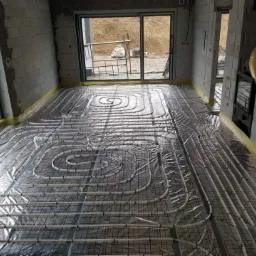 Instalacja ogrzewania podłogowego w surowym wnętrzu z widocznymi bloczkami betonowymi, rurami grzewczymi ułożonymi w spiralne wzory i folią izolacyjną, przygotowanie do wylewki.