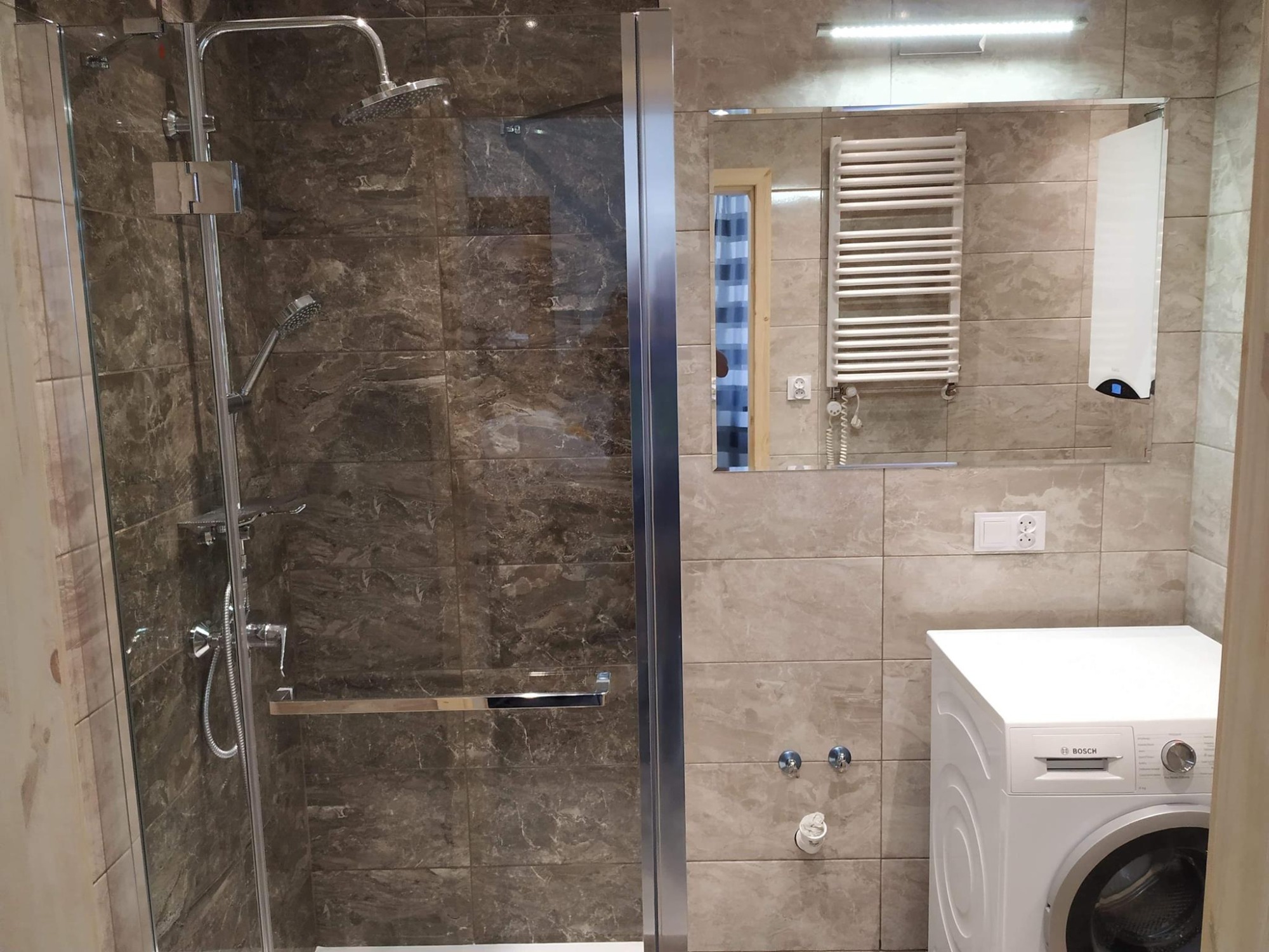 Nowoczesna łazienka z kabiną prysznicową wyłożoną marmurkowymi płytkami, widoczny grzejnik drabinkowy, lustro z oświetleniem LED oraz pralka Bosch