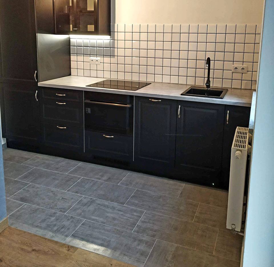 Ciemna zabudowa kuchenna z płytkami w kratkę na ścianie i szarą podłogą z płytek imitujących drewno, widoczny grzejnik.