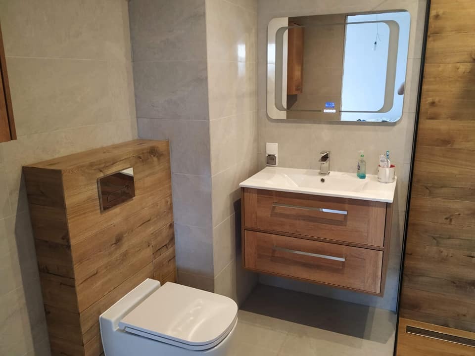 Nowoczesna łazienka z drewnianymi akcentami: wisząca szafka pod umywalką z dwiema szufladami, toaleta z zabudową w drewnie oraz lustro z podświetleniem LED.