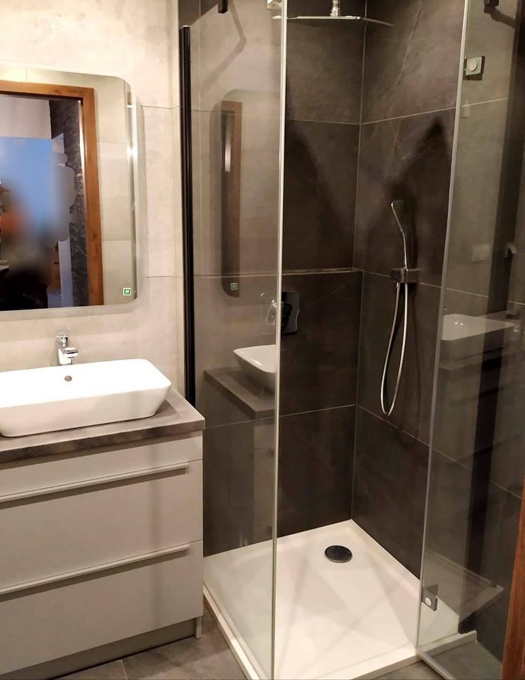 Nowoczesna łazienka z kabiną prysznicową typu walk-in, ciemnoszare płytki imitujące kamień, biały brodzik i umywalka nablatowa na szarej szafce, lustro w drewnianej ramie.
