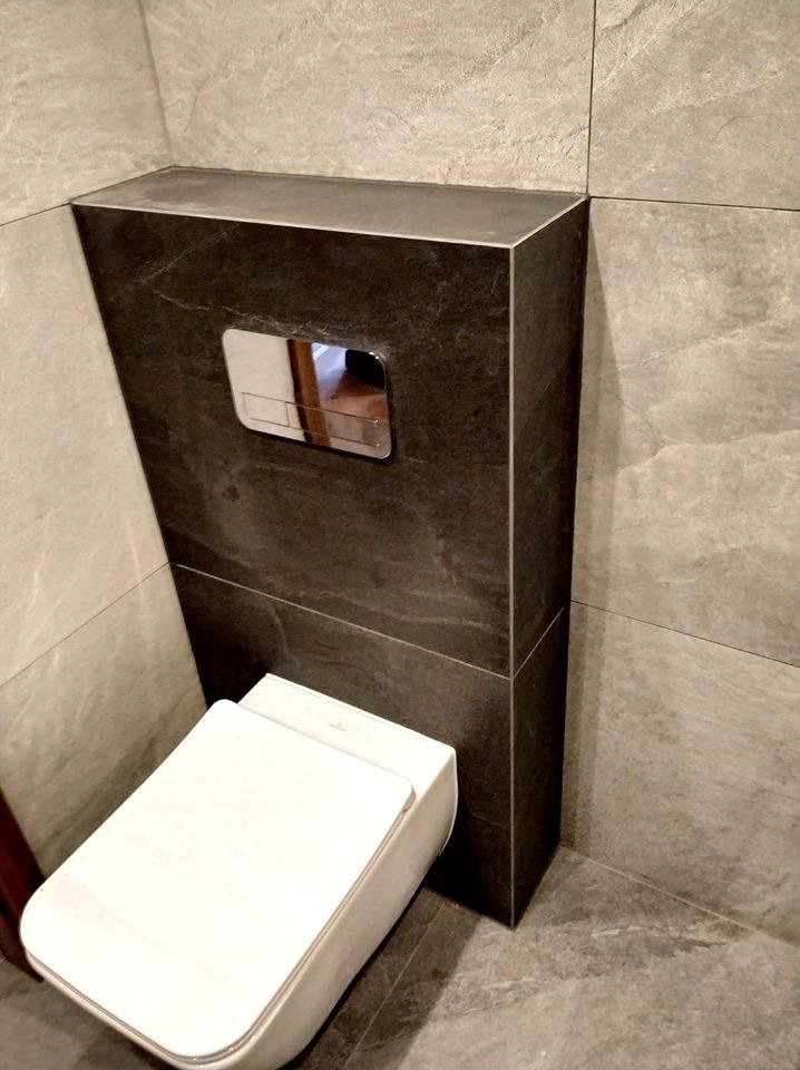 Nowoczesna toaleta podwieszana z czarną obudową z płytek imitujących kamień, chromowany przycisk spłukiwania, jasnoszare płytki na ścianach i podłodze.