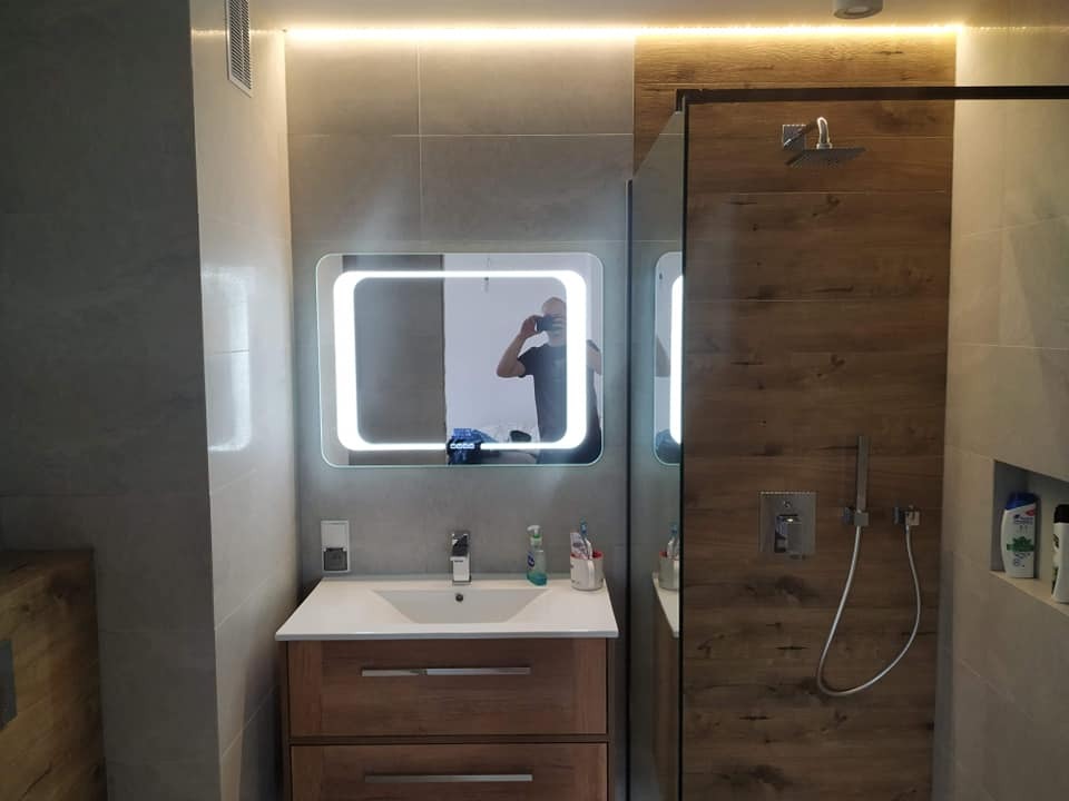 Nowoczesna łazienka z podświetlanym lustrem, kabiną prysznicową z drewnopodobnymi panelami i umywalką z szafką w odcieniach drewna.