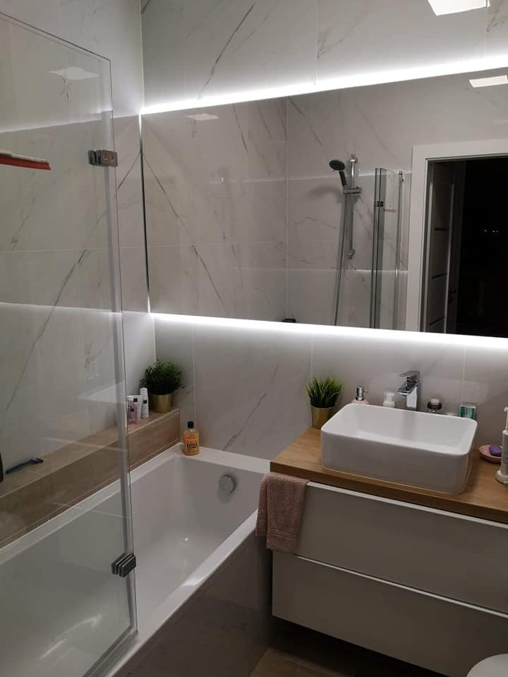 Nowoczesna łazienka z wanną za szklaną ścianką, umywalką nablatową na drewnianym blacie i podświetlanym lustrem na marmurowych płytkach.