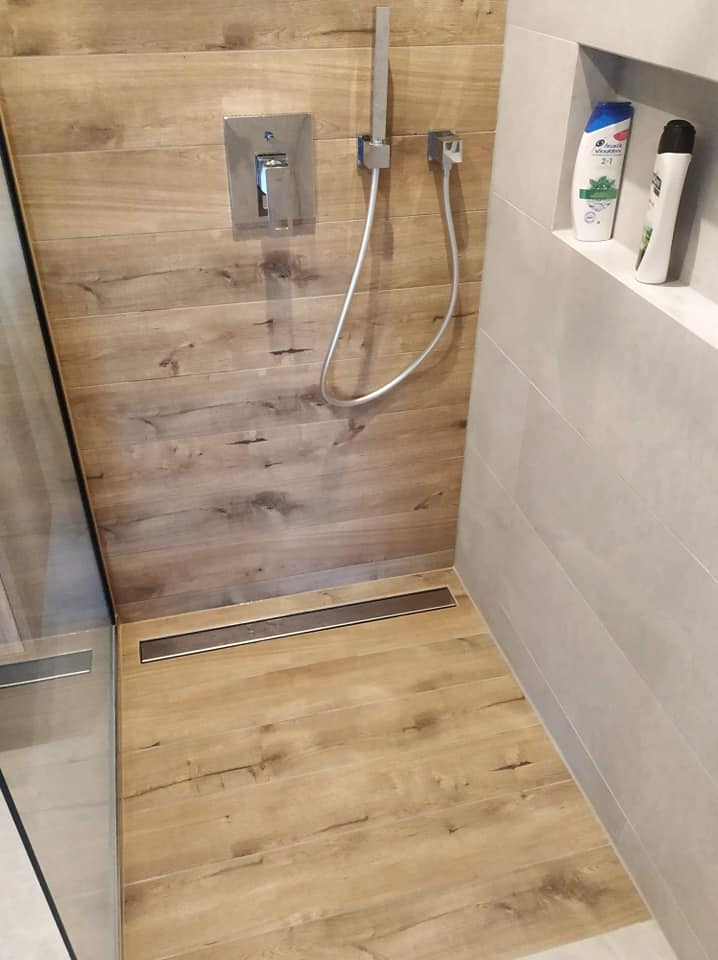 Kabina prysznicowa z odpływem liniowym w podłodze wyłożonej płytkami imitującymi drewno, ściany wykończone szarymi płytkami, wnęka z kosmetykami.