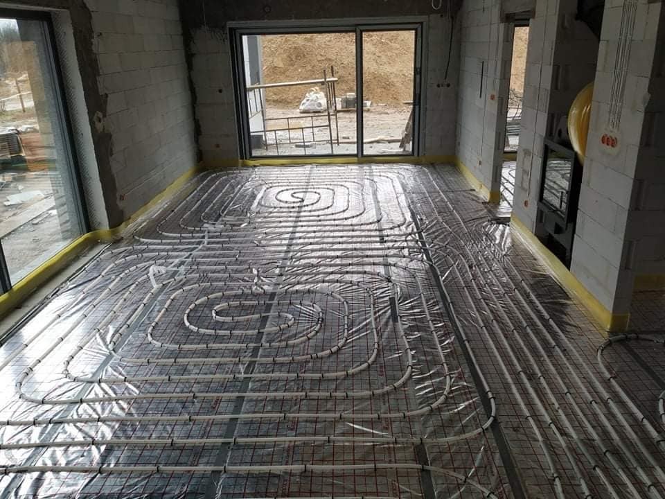 Instalacja ogrzewania podłogowego w surowym wnętrzu z widocznymi bloczkami betonowymi, rurami grzewczymi ułożonymi w spiralne wzory i folią izolacyjną, przygotowanie do wylewki.