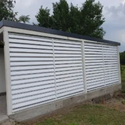 Nowoczesny garaż z białymi, poziomymi żaluzjami, betonową podstawą i ciemnym dachem na tle zachmurzonego nieba.