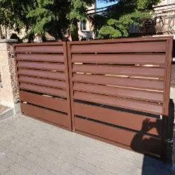 Nowoczesna, brązowa brama wjazdowa z poziomymi panelami, zamontowana na murowanych słupkach z dekoracyjnej cegły, na tle brukowanej nawierzchni i zieleni.