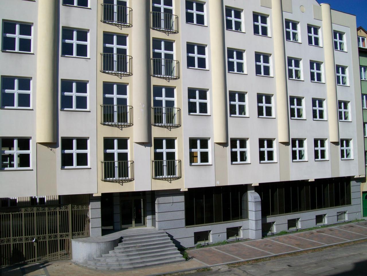 Fasada budynku mieszkalnego z balkonami i schodami wejściowymi, widok z zewnątrz w słoneczny dzień.