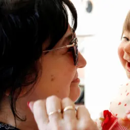 Uśmiechnięta dziewczynka w sukience z czerwoną kokardą patrzy na kobietę w okularach z dwoma złotymi pierścionkami na palcach.