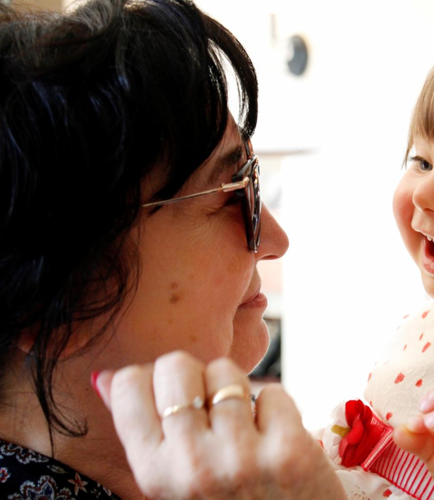 Uśmiechnięta dziewczynka w sukience z czerwoną kokardą patrzy na kobietę w okularach z dwoma złotymi pierścionkami na palcach.