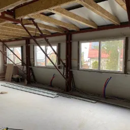 Poddasze w trakcie adaptacji: widoczne metalowe konstrukcje nośne, ocieplenie dachu płytami OSB, częściowo otynkowana ściana, okna z widokiem na domy, rury instalacyjne i profile pod płyty gipsowe...