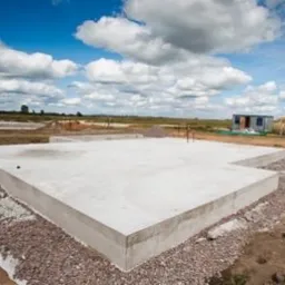 Surowy betonowy fundament domu jednorodzinnego na żwirowym podłożu, widoczne wystające pręty zbrojeniowe, w tle błękitne niebo z chmurami i kontener budowlany.