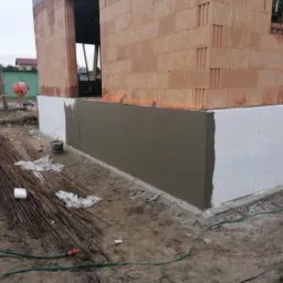 Ocieplony fundament budynku w trakcie prac wykończeniowych, widoczne materiały budowlane i mieszalnik w tle.