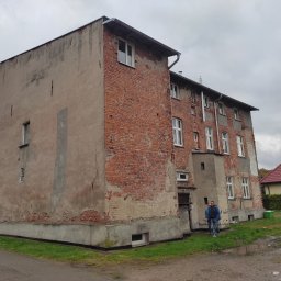 Starmit Marek Rek - Dwupiętrowy budynek z częściowo zniszczoną elewacją z czerwonej cegły i tynku, widoczny mężczyzna na zewnątrz. Budynek przed remontem.