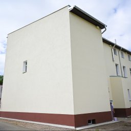 Starmit Marek Rek - Odnowiona elewacja budynku z kremowym tynkiem i brązowym cokołem, widoczna z perspektywy ulicy, z fragmentem znaku STOP i błękitnym niebem w tle.