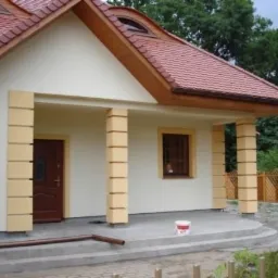 Jasny dom jednorodzinny z czerwonym dachem, kremową elewacją i żółtymi detalami kolumn, widoczny fragment rynny i wiaderko po farbie na ganku.