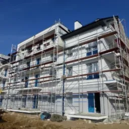 Wielopiętrowy budynek mieszkalny w trakcie ocieplania elewacji, widoczne rusztowania i częściowo ocieplona ściana, niebieskie niebo.