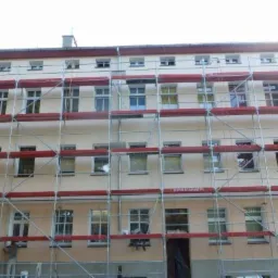 Wielopiętrowy budynek w trakcie malowania elewacji, z widocznymi rusztowaniami i pracownikami na nich.