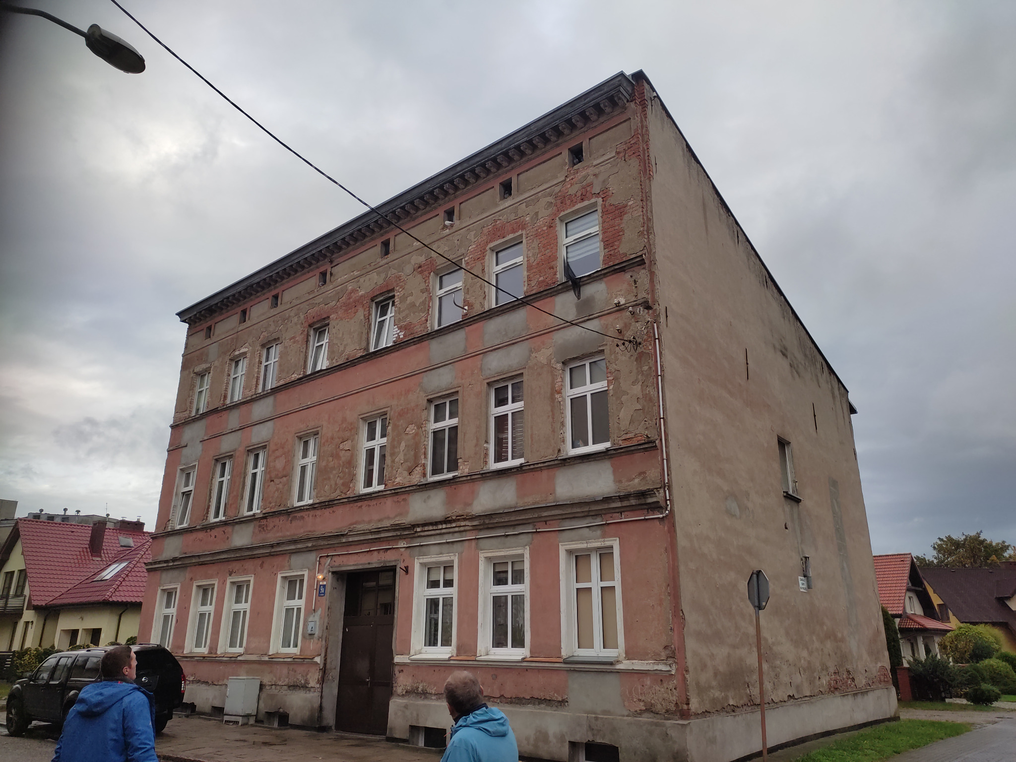 Trzypiętrowa kamienica z odpadającym tynkiem, widoczna z dołu pod kątem. Na pierwszym planie fragment chodnika i dwie osoby. W tle inne budynki i pochmurne niebo.