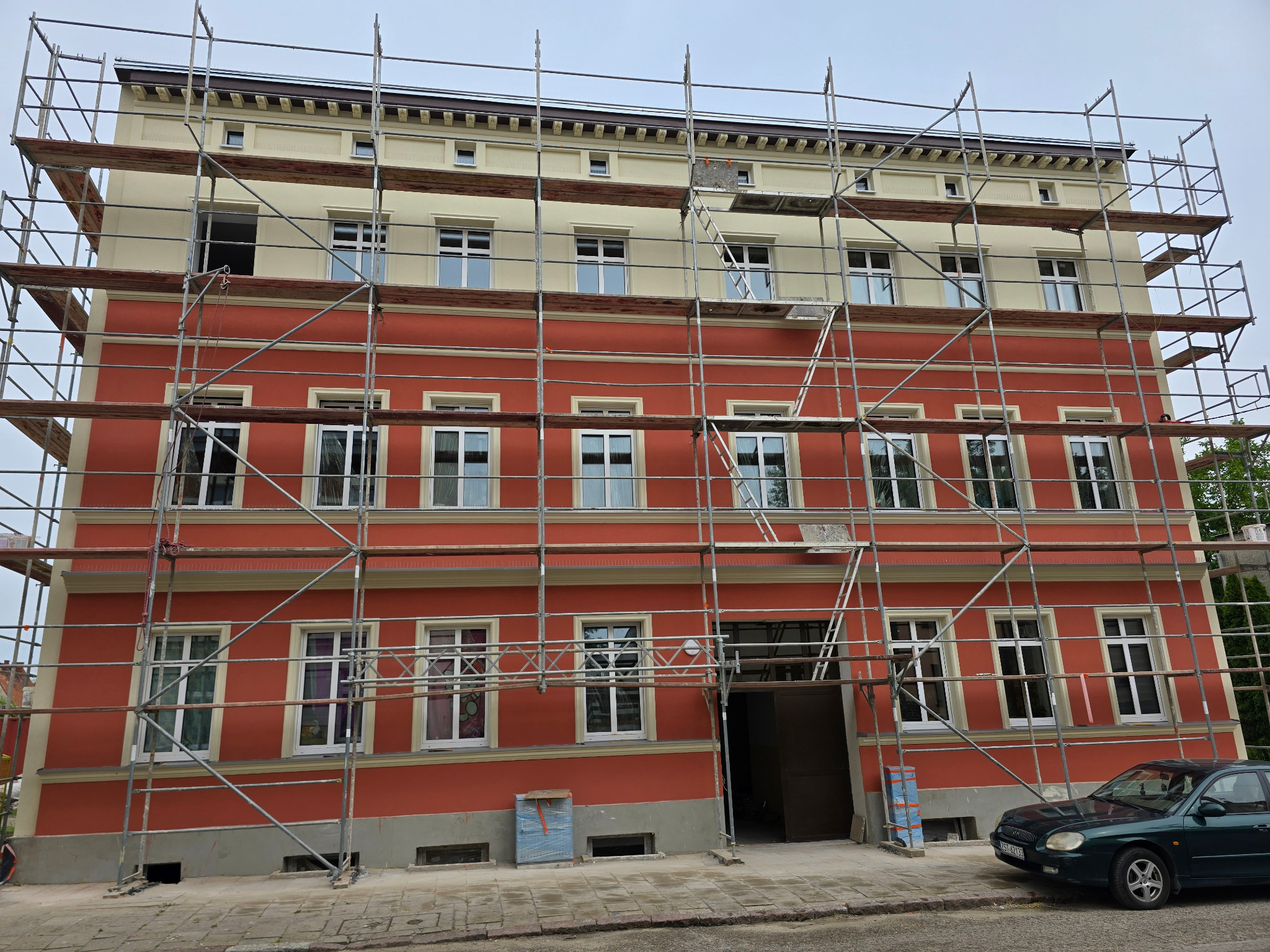 Czerwono-kremowa elewacja budynku w rusztowaniach. Widoczne okna z białymi ramami i fragment zielonego samochodu. Prace elewacyjne w toku.