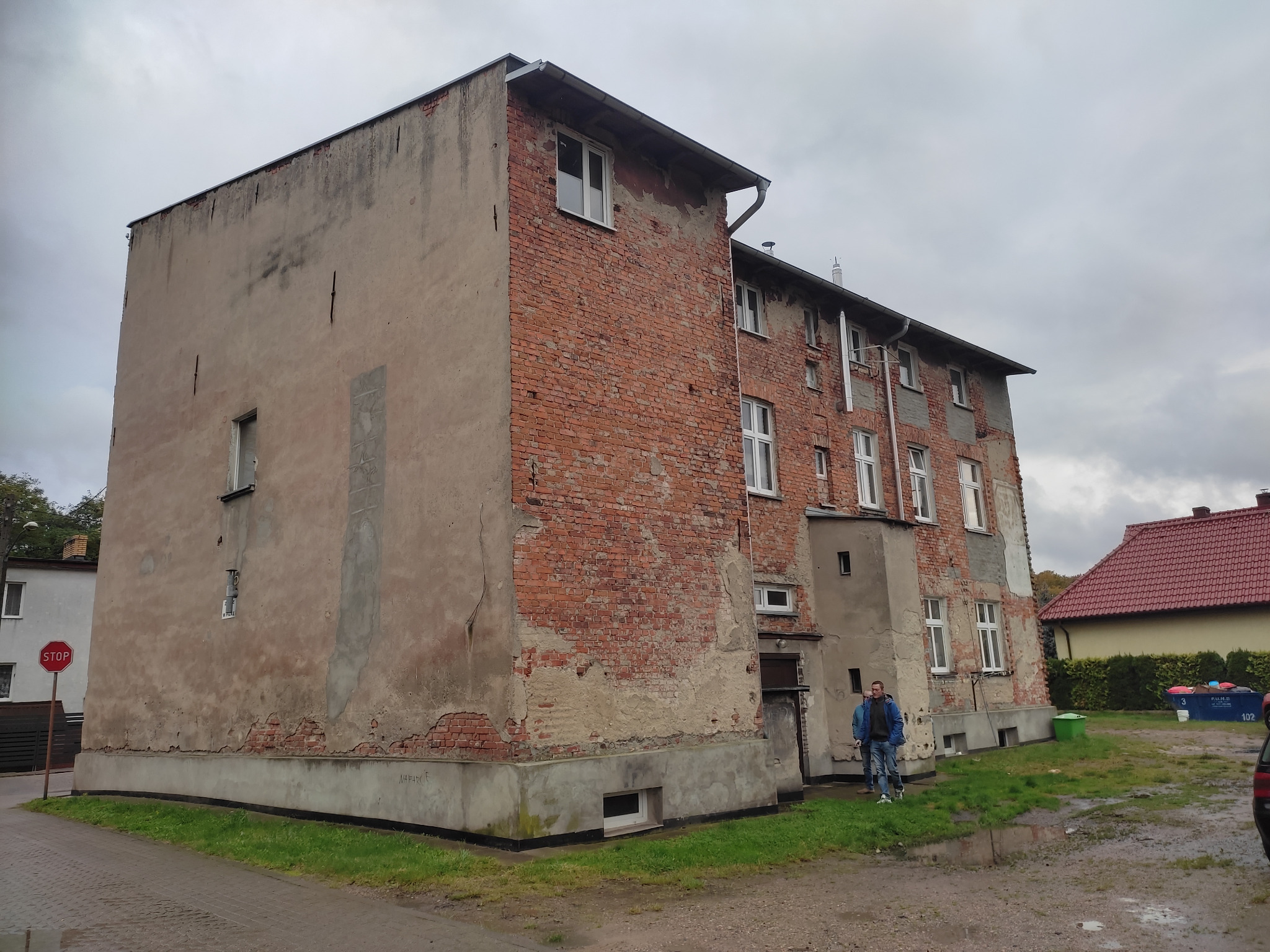 Dwupiętrowy budynek z częściowo zniszczoną elewacją z czerwonej cegły i tynku, widoczny mężczyzna na zewnątrz. Budynek przed remontem.