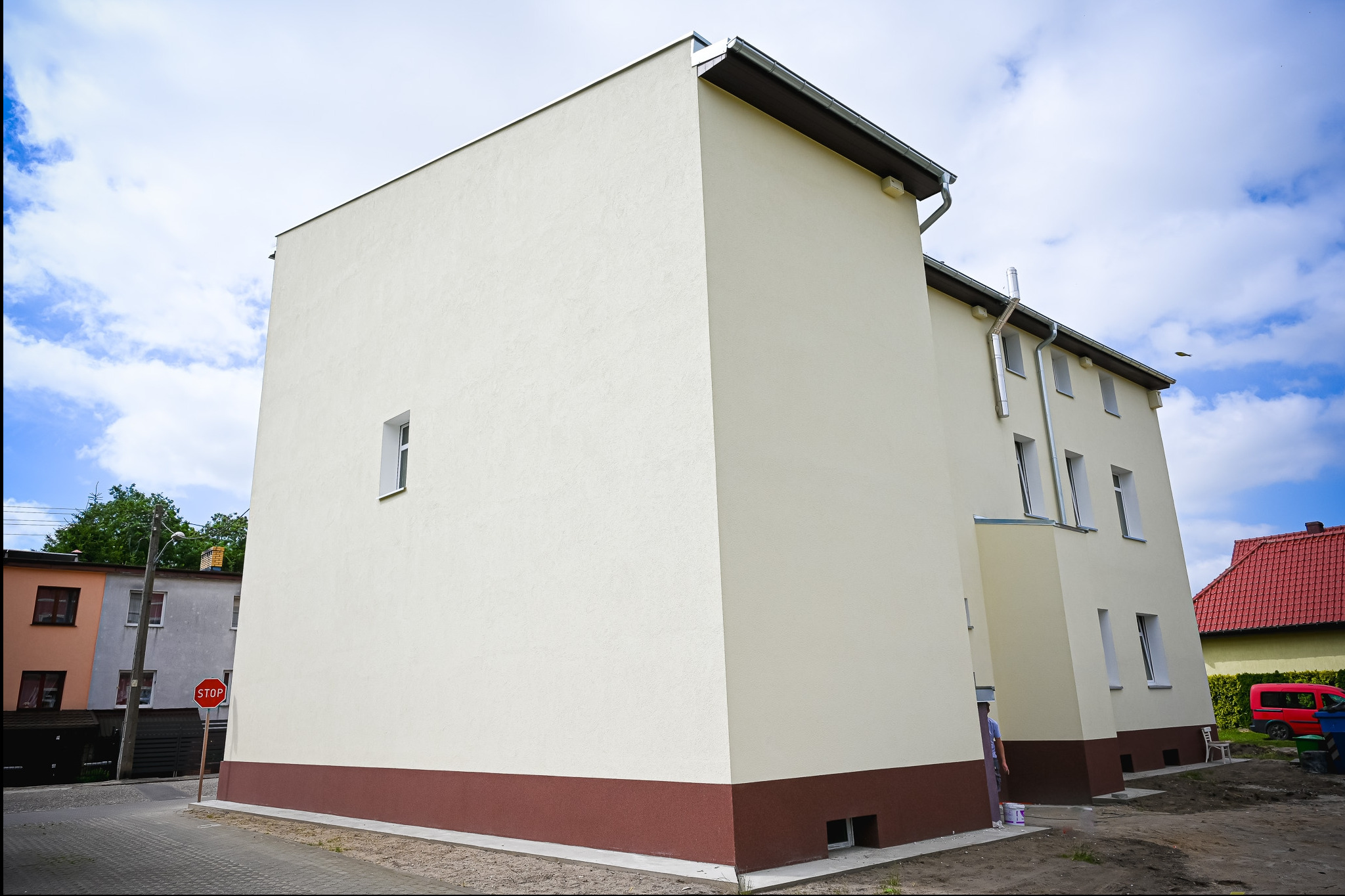 Odnowiona elewacja budynku z kremowym tynkiem i brązowym cokołem, widoczna z perspektywy ulicy, z fragmentem znaku STOP i błękitnym niebem w tle.