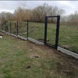 Montaż czarnego ogrodzenia panelowego z bramką na betonowej podmurówce, widoczne narzędzia i elementy montażowe na trawie, w tle drzewa bez liści.